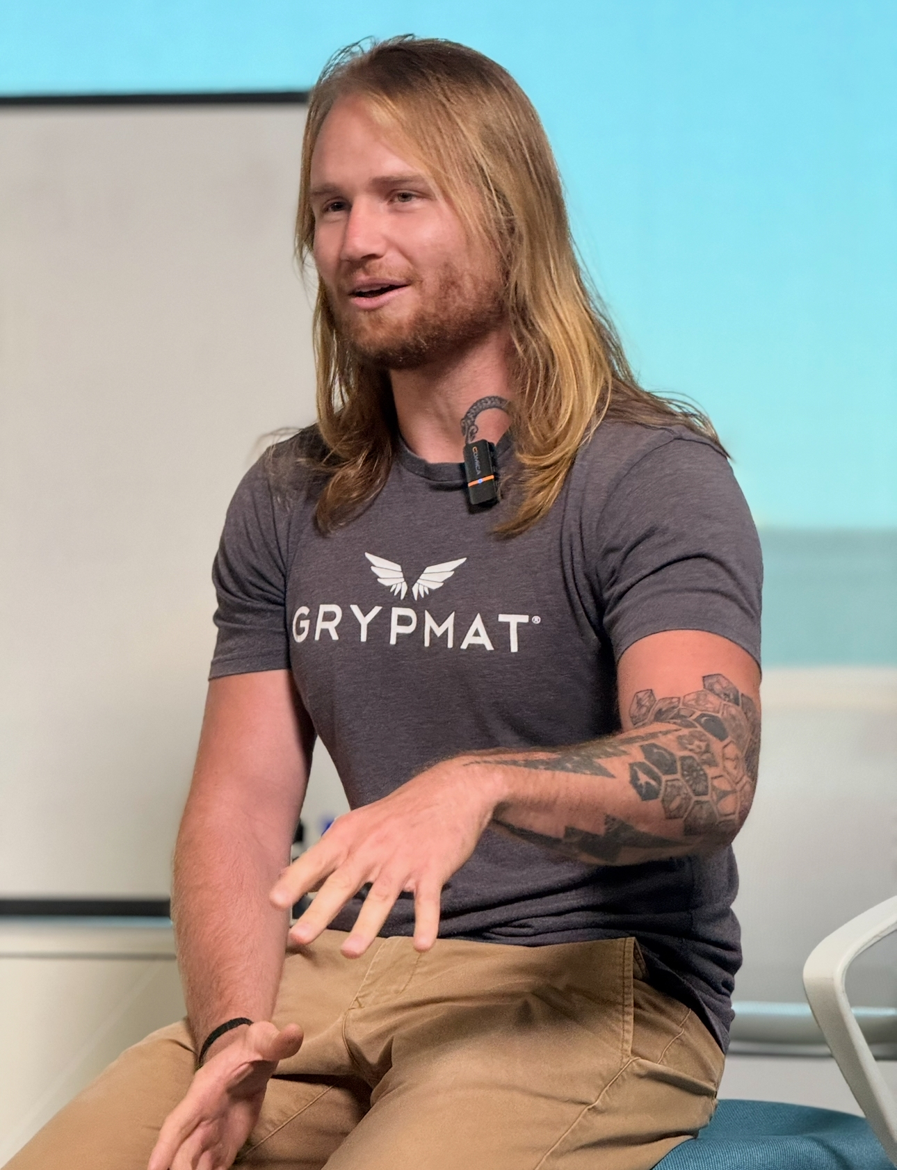 Tom Burden speaking in a Grypmat shirt, with long hair and visible tattoos